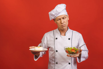 Picture of happy young senior chief cook in uniform standing isolated over red wall background, holding salad and cake. Diet concept.
