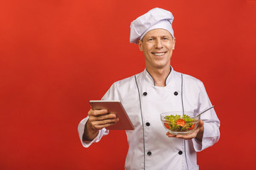 Picture of happy young senior chief cook in uniform standing isolated over red wall background, holding salad and tablet computer.