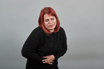 Young redhead fat lady in black sweater on grey background unhappy woman tilted body from stomach...