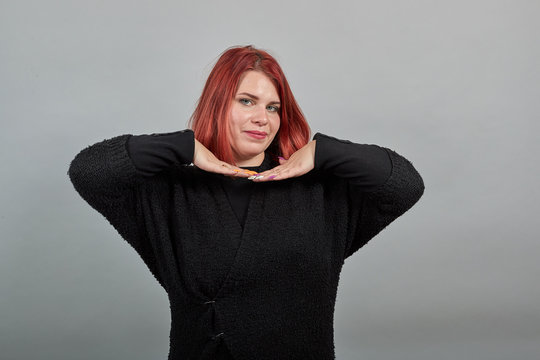 Young redhead fat lady in black sweater on grey background, cheerful woman holds her hands under head and dances with palms like in an Indian dance