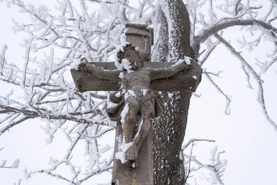 Beautiful Jesus Christ crucifix statue cover with snow, tree in background, cloudy winter day, religious meditation and contemplation concept