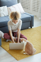 Pretty girl sitting on the floor and communicating online on laptop with cat sitting near by her in the room