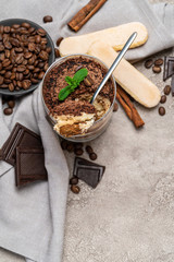 Classic tiramisu dessert in a glass cup, pieces of chocolate and savoiardi cookies on concrete background