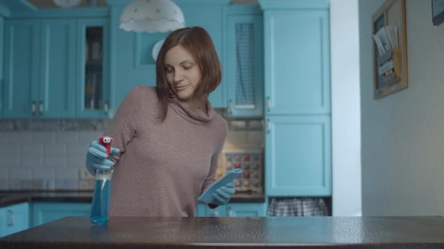 Young 30s female spraying detergent and wiping table surface with rag. Dancing and smiling woman making home clean up. 