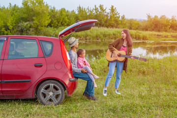 Family near the car outdoors and have fun. Happy father and little daughter sit in the trunk, and mother plays the guitar