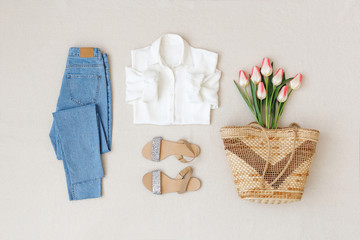 Blue jeans, white shirt, flat sandals, straw woven basket bag, bouquet of pink tulips flowers on beige background. Woman's casual spring summer outfit. Trendy women's clothes. Flat lay, top view.
