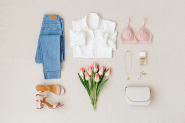 Blue jeans, white shirt, heeled sandals,  small clutch bag, bra, jewelry, bouquet of pink tulips on beige background. Woman's casual spring summer outfit. Trendy women's clothes. Flat lay, top view.