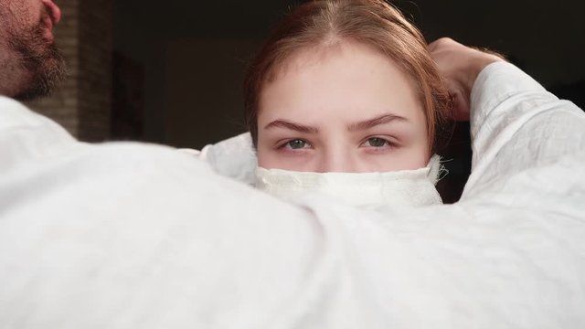 White man in white shirt ties medical face mask to face of white calm girl. BCU