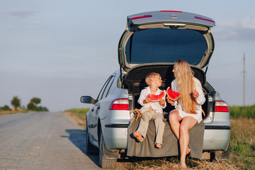 pretty blond hair woman with little blond son at sunset relaxing behind the car and eating watermelon. summer, travel, nature and fresh air in the countryside.