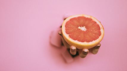 Female hand holding half of grapefruit through torn pink paper background. Fresh orange juice. Vegan, vegetarian concept. Banner with copy space.