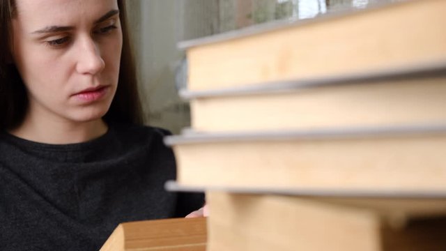 Young woman with heaps stack of books studying at home or library. Close up girl student sitting at the desk preparing for test exam with books writing essay coursework in notebook studying at home. 