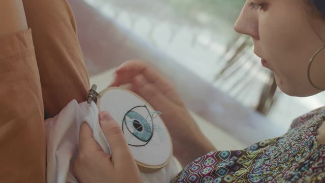 Woman embroidering eye pattern on white cloth lace-frame side view close up. Macro girl holding needle embroidery art outdoors natural light slow motion. Hobby technique equipment