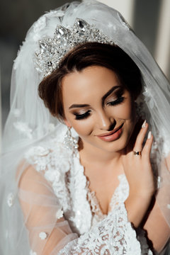 Bride with wedding makeup and hairstyle with crown. Bride touch your earrings. Fashion bride. Attractive bride in white robe in wedding morning.