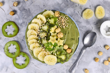 Green fruit kale smoothie bowl topped with star shaped banana, kiwi, hazelnut and puffed quinoa grain on gray background