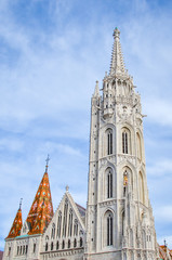 The spire of the famous Matthias Church in Budapest, Hungary. Roman Catholic church built in the Gothic style. Orange colored tile roof. Blue sky and white clouds above. Vertical photo