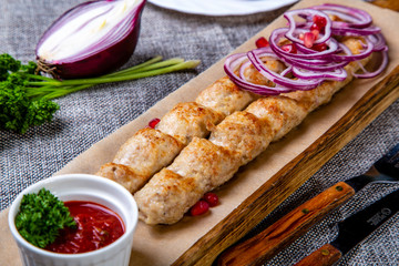 Lula-kebab of chicken with onions and hot red sauce. Served on a wooden tray. Background-grey linen. Around the cradle are spread onions, parsley, pomegranate and a knife and fork