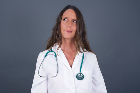 Photo of amazed puzzled young caucasian doctor female curves lips and has worried look, sees something awful in front, isolated on white background, wearing medical uniform.
