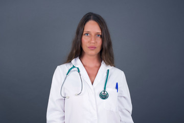 Waist up shot of joyful doctor girl wearing medical uniform looking to the camera, thinking about something. Both arms down, neutral facial expression wondering about something happening.