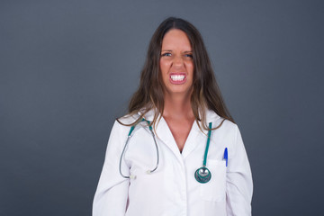Mad crazy woman clenches teeth angrily, being annoyed with coming noise, dressed in fashionable clothes, isolated over gray background. Negative feeling concept.