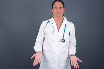 Puzzled and clueless young doctor woman with arms out, shrugging her shoulders, saying: who cares, so what, I don't know.