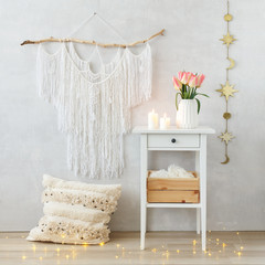 Spring boho home interior decor: macrame wall hanging decoration, white bedside table, vase, pink tulips flowers, lighted candles, pillow, led garland glowing lights. Light cozy modern stylish room. © NesolenayaAleksandra