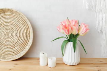 White ceramic vase with bouquet of pink tulips flowers, wicker tray and candles on a wooden table or shelf on a background of light gray wall. Stylish feminine spring home interior decor.