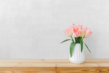 White geometric ceramic vase with bouquet of pink tulips flowers on a wooden table or shelf on a background of light gray wall. Simple minimal stylish spring home interior decor. Space for text.
