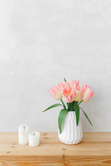 White geometric ceramic vase with bouquet of pink tulips flowers and candles on a wooden table or shelf on a background of light gray wall. Simple spring home interior decor.