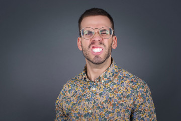 Obraz premium Mad crazy man clenches teeth angrily, being annoyed with coming noise, dressed in fashionable clothes, isolated over gray background. Negative feeling concept.