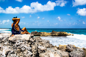 Young sexy woman sitting on the rocks in bikini holds her hat and looks at the waves of the tropical sea on the horizon, paradise and summer, happy and freedom, horizontal image