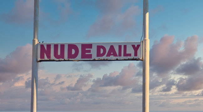 An old rusty, beat up, sign saying Nude Daily on a blue sky