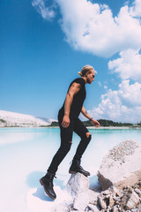 Curly blond man posing at the clear beach. Dressed all black. Awesome background, clear water and sky. Trendy, tropical and attractive man. Summer time