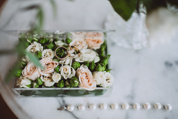 Wedding rings on a floral surface with roses and green blossom. Transparent box with jewelry and flowers, lying on a white marble table.
