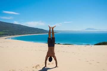 Male guy with a muscled back doing a handstand in a beautiful dune next to the atlantic in Tarifa,...