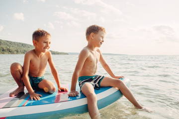 Two smiling Caucasian boys kids sitting on paddle sup surfboard in water. Children friends talking laughing. Modern outdoor summer activity. Aquatic seasonal sport hobby. Happy childhood lifestyle.