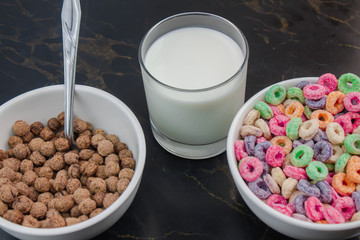 Fruit cereal and chocolate cereal in white cups next to glass of milk in minimalist black table