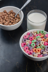 Fruit cereal and chocolate cereal in white cups next to glass of milk in minimalist black table
