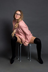 Obraz na płótnie Canvas a blonde with long straight hair in a braid, in a pink short dress, posing in a Studio on a gray background, Sitting on a stool, legs wide apart, in black leggings.