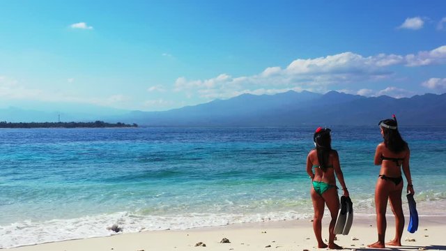 Snorkeling in the tropical ocean, two sexy ladies in the bikinis with snorkeling masks and fins talking on the sandy beach