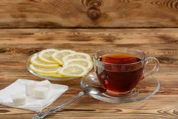 A glass cup of tea on a wooden table with sugar and slices of lemon in a plate.