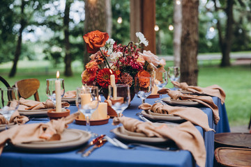 festive tables with tablecloths made of textiles are decorated with flower arrangements, on the tables are candles, plates with napkins, glasses, cutlery, a wedding in the forest, banquet, holiday