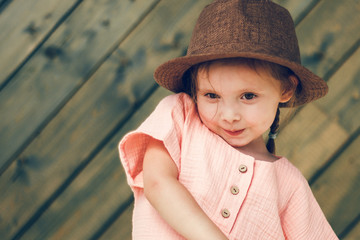 Portrait of happy cute little girl in rural style brown hat and muslin clothes on green wooden...