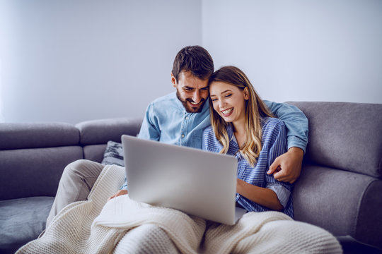 Cheerful cute caucasian couple sitting on sofa covered with blanket and using laptop for internet surf. Living room interior.