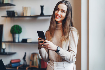 Female office worker standing at window, using smartphone for communication. Girl with smartphone in hand, surfing the Internet, pretty woman looking at camera. Digital tools.