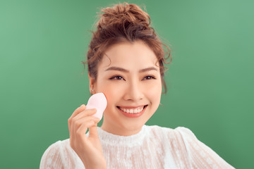 Beauty portrait of a lovely young asian woman standing isolated over pink background, using makeup sponge