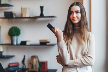 Beautiful woman with long hair standing by window, using smartphone to communicate. Girl with celltphone in hand surfing Internet. Office worker in good mood during break in work. Digital tools.