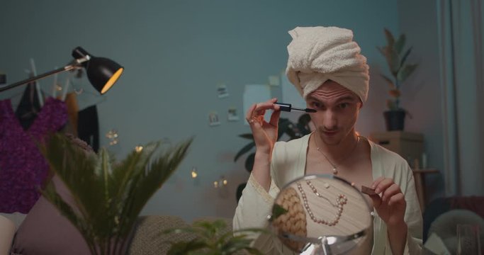 Young man transvestite in bath robe getting ready for date and doing make up. Man transgender using mascara for eyelashes and eating piece of chocolate while looking in small mirror.