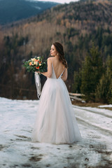 Fototapeta na wymiar cute bride in a white dress with a neckline on her back holds a bouquet on a background of a winter forest.