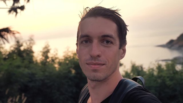 Portrait of man tourist making selfie or a video call using smartphone outdoors on background of evening tropical island sunset