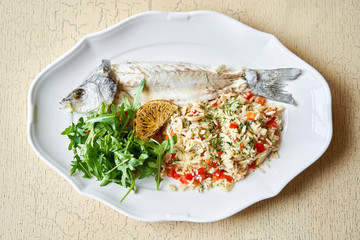 Grilled sea bass. Salad and rice with fresh vegetables. Serving on a white plate on wooden table. Restaurant menu.